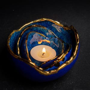 May include: A blue ceramic candle holder with a gold rim, holding a lit white candle. The candle holder has a layered design, with the candle nestled in the center. The gold rim adds a touch of elegance to the piece.