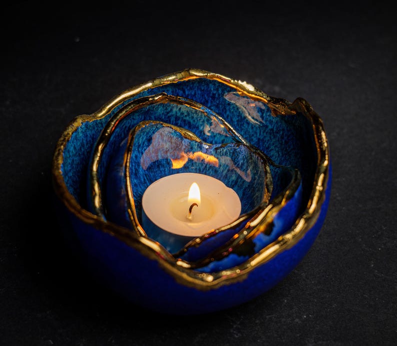 May include: A blue ceramic candle holder with a gold rim, holding a lit white candle. The candle holder has a layered design, with the candle nestled in the center. The gold rim adds a touch of elegance to the piece.
