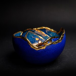 May include: A blue ceramic candle holder with gold trim. The candle holder has a textured surface and a lit candle inside, creating a warm glow. The gold trim adds a touch of elegance to the design.