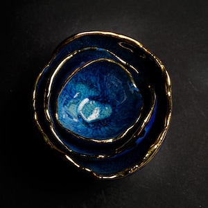 May include: A set of three nested ceramic bowls in varying sizes. The bowls are a deep blue with a glossy finish and gold-colored rims. The bowls are arranged in a nested configuration, with the smallest bowl at the center.