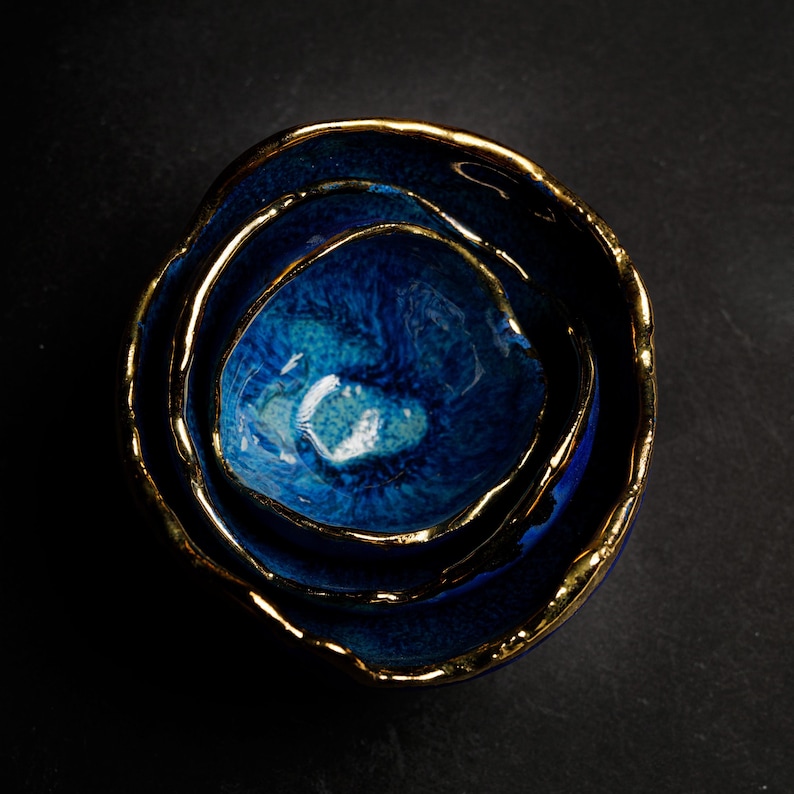 May include: A set of three nested ceramic bowls in varying sizes. The bowls are a deep blue with a glossy finish and gold-colored rims. The bowls are arranged in a nested configuration, with the smallest bowl at the center.