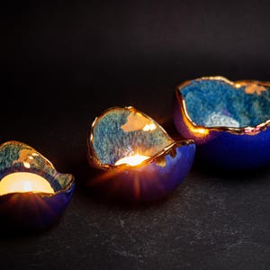 May include: Three ceramic candle holders in varying sizes, each with a deep blue exterior and a gold-colored rim. The interior has a teal and gold pattern. Lit candles glow inside the holders, creating a warm ambiance.