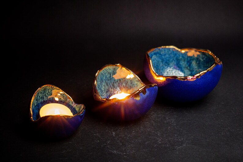 May include: Three ceramic candle holders in varying sizes, each with a deep blue exterior and a gold-colored rim. The interior has a teal and gold pattern. Lit candles glow inside the holders, creating a warm ambiance.