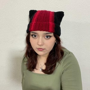 Op de afbeelding: Een rode en zwarte gebreide beanie met kattenoren. De beanie is gemaakt van zacht, warm garen en heeft een comfortabele pasvorm.