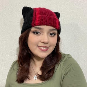 Op de afbeelding: Een rood en zwart gebreide beanie met kattenoren. De beanie heeft een gestreept patroon en wordt gedragen door een persoon met lang donker haar.