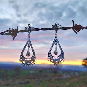 Könnte beinhalten: Ein Paar silberne Ohrringe mit filigranen Designs. Die Ohrringe hängen an einem Stacheldrahtzaun vor einem Sonnenuntergang.