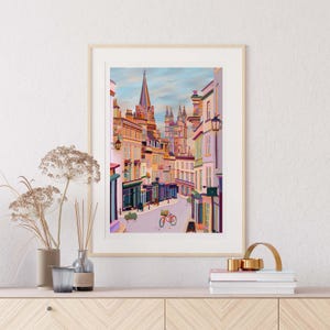 May include: A framed illustration of a narrow street in a city with colourful buildings. The street is lined with shops and a bicycle is parked in the middle of the street. The buildings are in a variety of pastel colours and the street is paved with cobblestones.