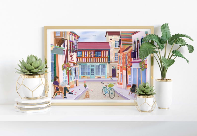 May include: A framed print of a colorful street scene with buildings, a bicycle, and a person reading. The print is displayed on a white shelf with decorative plants in white and gold pots.
