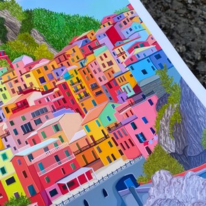 May include: A colourful illustration of a hillside village with buildings in shades of pink, yellow, blue, green, and red. The buildings are stacked on top of each other, creating a sense of depth and perspective. The village is nestled against a rocky cliffside, with lush green foliage growing on the slopes.