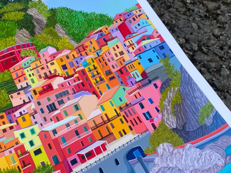 May include: A colourful illustration of a hillside village with buildings in shades of pink, yellow, blue, green, and red. The buildings are stacked on top of each other, creating a sense of depth and perspective. The village is nestled against a rocky cliffside, with lush green foliage growing on the slopes.