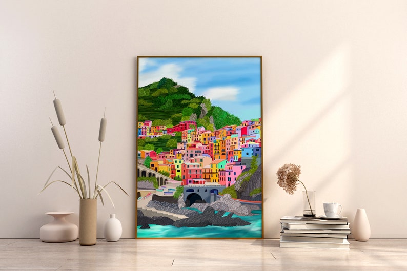 May include: A colourful painting of a seaside town with brightly coloured buildings on a hillside. The town is nestled between a green hill and the blue ocean. The painting is framed in a gold frame.