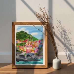 May include: A framed print of a colourful illustration of a seaside town with buildings on a hillside. The town is painted in shades of pink, yellow, blue, green, and orange. The water is a bright blue and there are rocks and trees in the foreground.