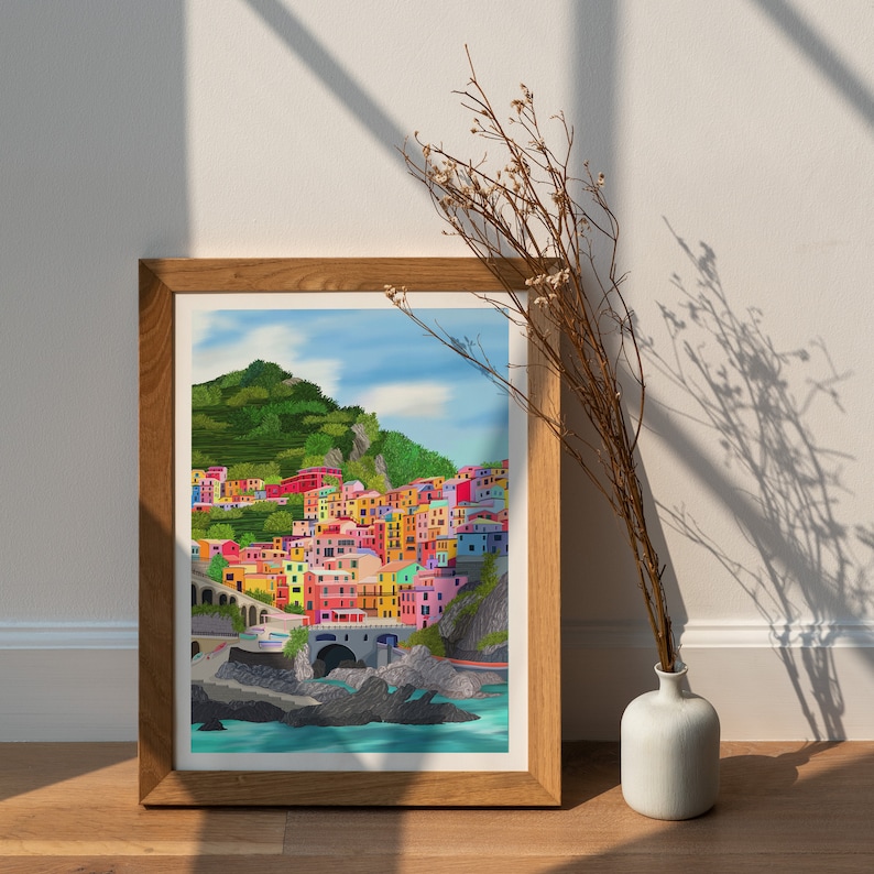 May include: A framed print of a colourful illustration of a seaside town with buildings on a hillside. The town is painted in shades of pink, yellow, blue, green, and orange. The water is a bright blue and there are rocks and trees in the foreground.