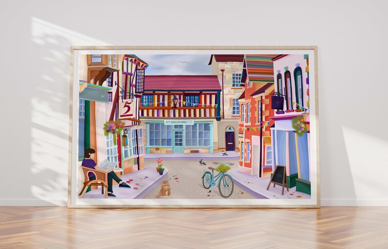 May include: A framed art print depicting a quaint European street scene. The artwork features colorful buildings, a bicycle, and a person reading a newspaper. The buildings have red, orange, and blue facades. The print is displayed on a wooden floor.
