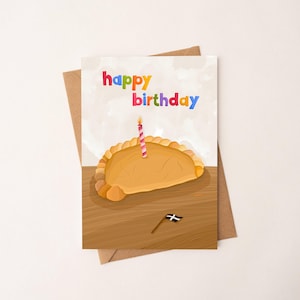 May include: A birthday card with a cartoon illustration of a pasty with a lit candle. The words "happy birthday" are written in colourful letters. A small flag is also included in the illustration.