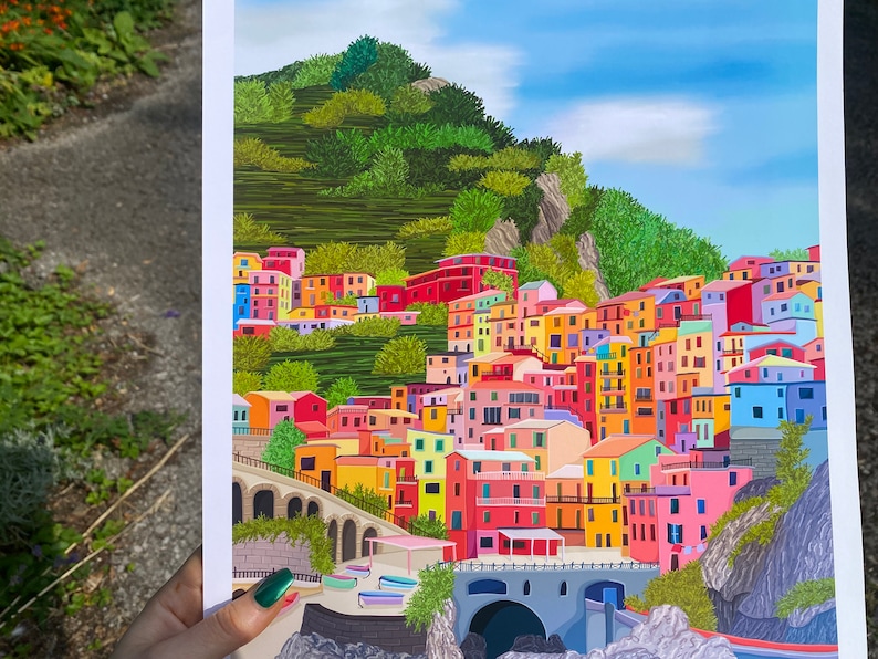 May include: A colourful illustration of a hillside village with buildings in shades of pink, yellow, blue, green, and red. The village is nestled against a green hillside with a blue sky above.