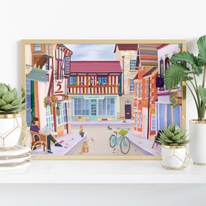 May include: A framed art print depicting a colorful street scene with buildings, a bicycle, and a person reading. The print is displayed on a white shelf with potted plants in white and gold geometric planters.