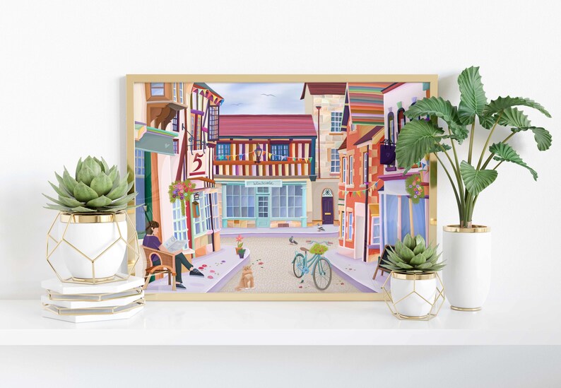 May include: A framed art print depicting a colorful street scene with buildings, a bicycle, and a person reading. The print is displayed on a white shelf with potted plants in white and gold geometric planters.
