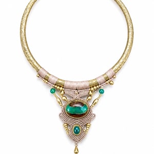 May include: A gold-toned metal torque necklace featuring intricate etched patterns, beige macramé cord detailing, and a central oval green stone. The design includes smaller green beads, gold-toned metal accents, and a small dangling gold-toned drop.