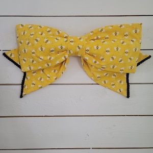 May include: A large yellow bow with a black trim and a pattern of small bees. The bow is made of fabric and is tied in a large, symmetrical knot.