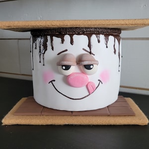 May include: A white ceramic marshmallow with a chocolate drizzle and googly eyes, sitting on a brown graham cracker base. The marshmallow has a pink nose and a brown smiling mouth with a red tongue sticking out.