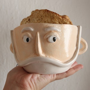 May include: A novelty ceramic mug in the shape of a face, painted in a light peach hue with white details. The mug features a handle and contains a slice of toasted bread. The face has detailed eyes and a mustache.