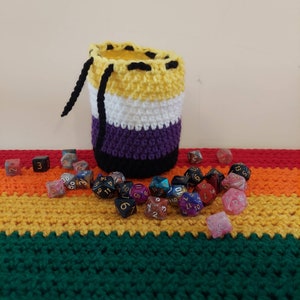 May include: A crocheted dice bag with a yellow, white, purple, and black color scheme, representing the non-binary pride flag. The bag is filled with colorful dice, some with numbers and some with symbols.