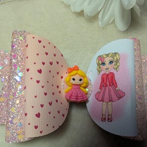 May include: A pink hair bow with glitter detailing and a heart pattern. The bow features a cartoon doll in a pink dress and a yellow bow. This decorative bow is a hair accessory.
