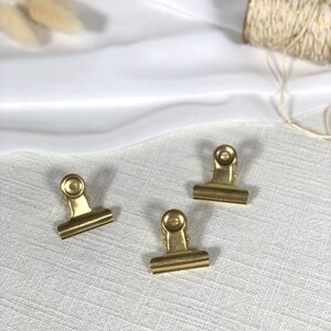 Könnte beinhalten: Drei goldene Büroklammern auf einer weißen, strukturierten Oberfläche. Die Klammern sind aus Metall und haben einen Federmechanismus.