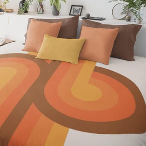 May include: A bed dressed with a duvet cover in a retro style, showcasing a circular design in brown, orange, and yellow. The bed is complemented by pillows in various shades of brown, orange, and yellow, creating a warm atmosphere.