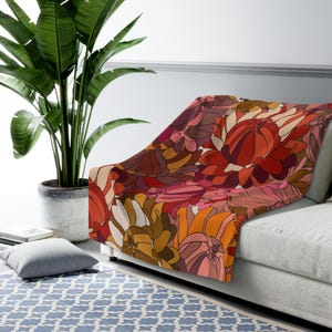 May include: A soft, plush throw blanket with a retro floral pattern in shades of red, orange, pink, and brown. The blanket is folded and draped over a white couch.