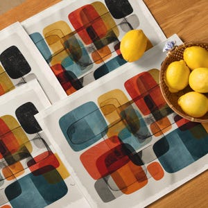May include: A set of white placemats with an abstract design of overlapping shapes in orange, yellow, blue, and black. A woven basket filled with yellow lemons sits on the table, next to the placemats. The placemats are on a wooden surface.