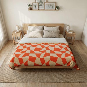 May include: A bed blanket featuring a repeating wavy, distorted checkerboard pattern in vibrant orange and light beige. The blanket is draped over a bed with four neutral-colored pillows, positioned within a bedroom setting with wooden furniture.