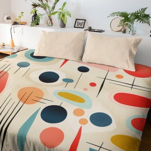 May include: A bed with a cream-colored duvet cover featuring an abstract design of colorful shapes. The design includes circles, ovals, and lines in shades of red, blue, yellow, and orange. Two cream-colored pillows rest on top of the duvet.
