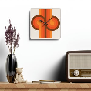 May include: Square wall clock with a retro design. The clock face features a cream background with a brown, orange, and yellow graphic. Black clock hands indicate the time. The clock is displayed on a wooden shelf with a vase, a teddy bear, and a vintage radio.