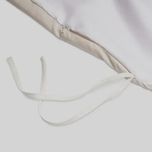 May include: Close-up of a white duvet cover with a beige border and a zipper closure. White fabric ties are visible at the corner. The bedding is made of a soft, smooth material.