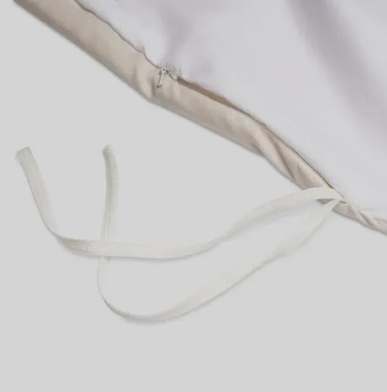 May include: Close-up of a white duvet cover with a beige border and a zipper closure. White fabric ties are visible at the corner. The bedding is made of a soft, smooth material.