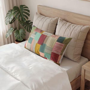 Peut inclure: Un oreiller rectangulaire présentant un motif patchwork composé de blocs de différentes couleurs, incluant des nuances de rouge, vert, jaune et crème, avec de fines lignes verticales et horizontales. Il est placé devant deux oreillers matelassés beiges.