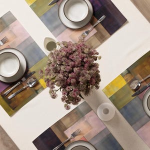 May include: Overhead view of a dining table set for four. Each place setting includes a white plate and bowl with a dark rim, silverware, and a colorful placemat. A centerpiece of purple flowers sits in the middle. The table is covered with a white tablecloth.