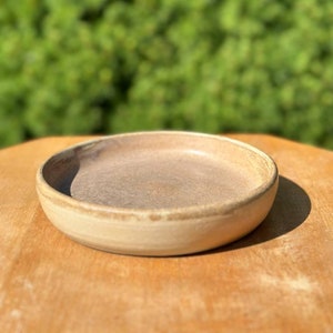Puede incluir: Un pequeño plato redondo de cerámica beige con un borde ligeramente elevado. El plato está vacío y tiene una textura sutilmente moteada.