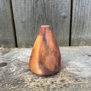 May include: A small, triangular ceramic vase with a brown, orange and red glaze. The vase has a narrow opening at the top and a wide base.