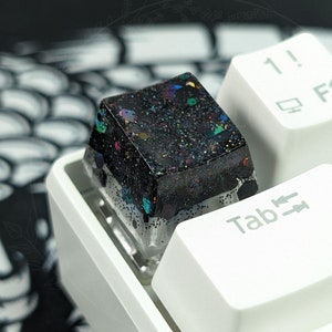 May include: A black and white keyboard with a custom keycap. The keycap is made of resin and has a glittery finish. The keycap is labeled "Tab" and has arrows pointing up and down.