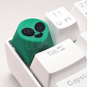 Puede incluir: Un tapón de tecla verde con un diseño de cara de extraterrestre negro. El tapón de tecla está en un teclado blanco.