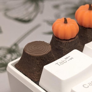 Peut inclure: Un clavier blanc avec des touches en forme de citrouille brunes et orange. Les touches sont en forme de souches d'arbres avec des citrouilles sur le dessus. Le clavier a des touches étiquetées "Tab", "Q", "W", "A" et "Caps Lock".