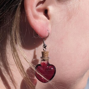 May include: A pair of heart-shaped glass bottle earrings with a cork stopper and a dark red liquid inside. The earrings are hanging from silver hooks.