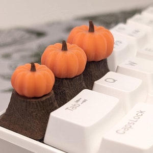 May include: Three miniature orange pumpkins with brown stems are placed on top of brown, tree stump-shaped keycaps. The keycaps are on a white keyboard with white keys. The keys are labeled "Tab", "Q", "W", "E", "R", "T", "Y", "U", "I", "O", "P", "A", "S", "D", "F", "G", "H", "J", "K", "L", "Z", "X", "C", "V", "B", "N", "M", ",", ".", "/", "Caps Lock".