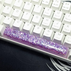 May include: A white mechanical keyboard with a purple and iridescent glitter keyboard keycap. The keyboard is on a black mousepad.