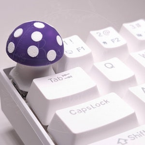 May include: A white keyboard with a purple and white mushroom keycap on the Tab key. The keyboard has keys labeled Tab, Caps Lock, and Shift.