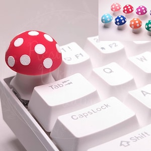 May include: Red and white mushroom-shaped keycap for a computer keyboard. The keycap is made of silicone and has a white polka dot pattern on a red background.