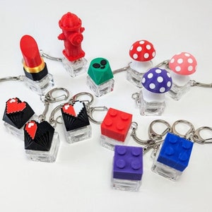 May include: A set of nine keychains with different charms, including a red fire hydrant, a red lipstick, a red and white heart, a black and red heart, a green alien, a red mushroom, a white mushroom with purple polka dots, a pink mushroom, a red Lego brick, a blue Lego brick, and a purple Lego brick. Each charm is attached to a clear acrylic keychain.